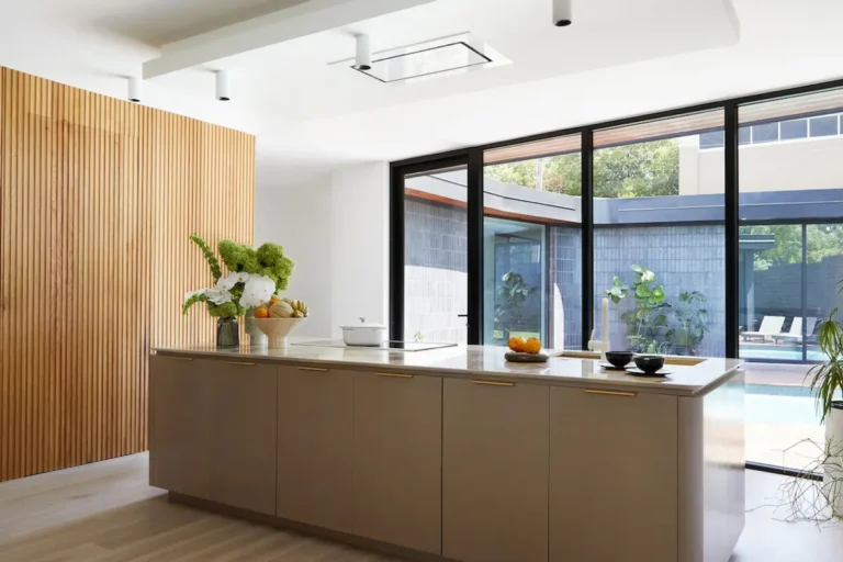 Большой современный коричневый кухонный остров с мойкой и варочной поверхностью на белой кухне со стеклянными окнами от пола до потолка